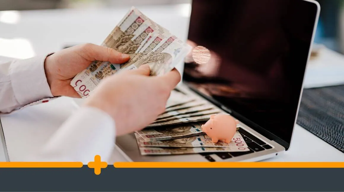 Ręce trzymające gotówkę nad komputerem z miniaturową skarbonką